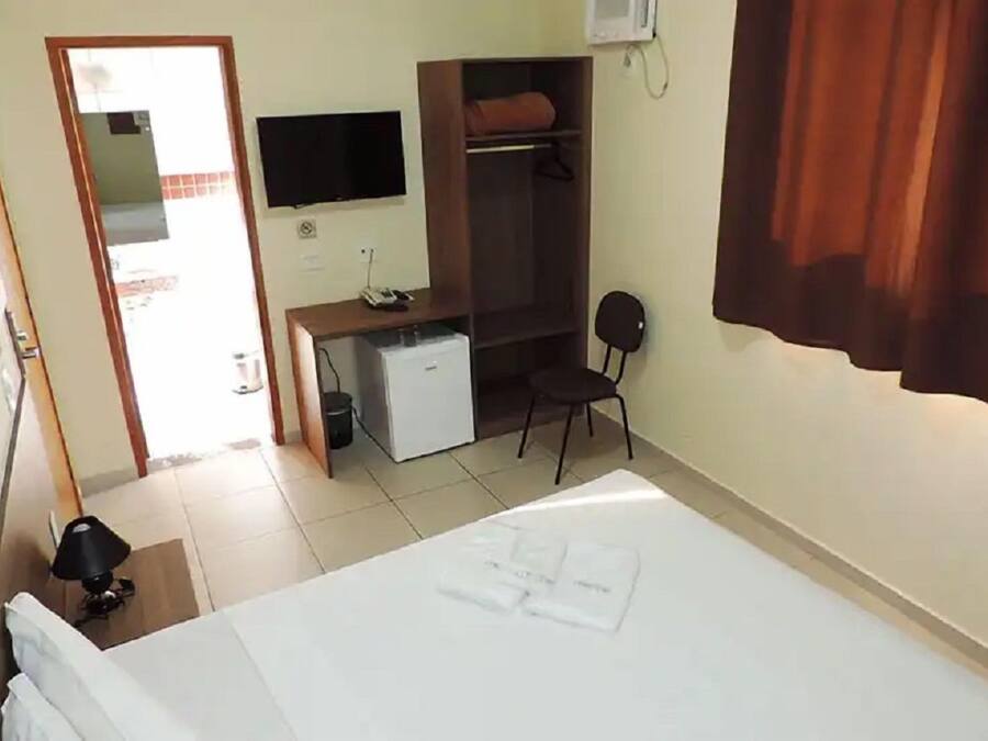 A hotel room with a bed, a desk with a computer, a small fridge, a chair, and a TV mounted on the wall.
