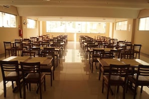 Uma sala de jantar espaçosa com várias mesas e cadeiras, janelas amplas e um extintor de incêndio na parede.