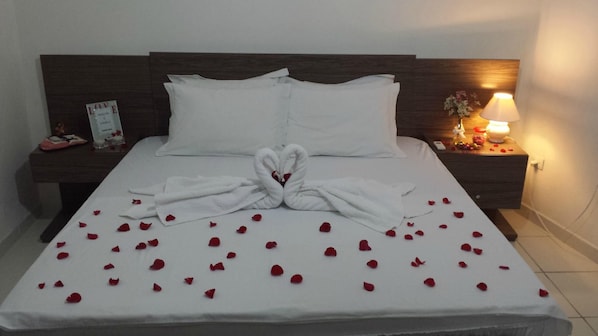 Una cama bien hecha con ropa de cama blanca y un arreglo de toalla con forma de cisne, rodeada de pétalos de rosa esparcidos.