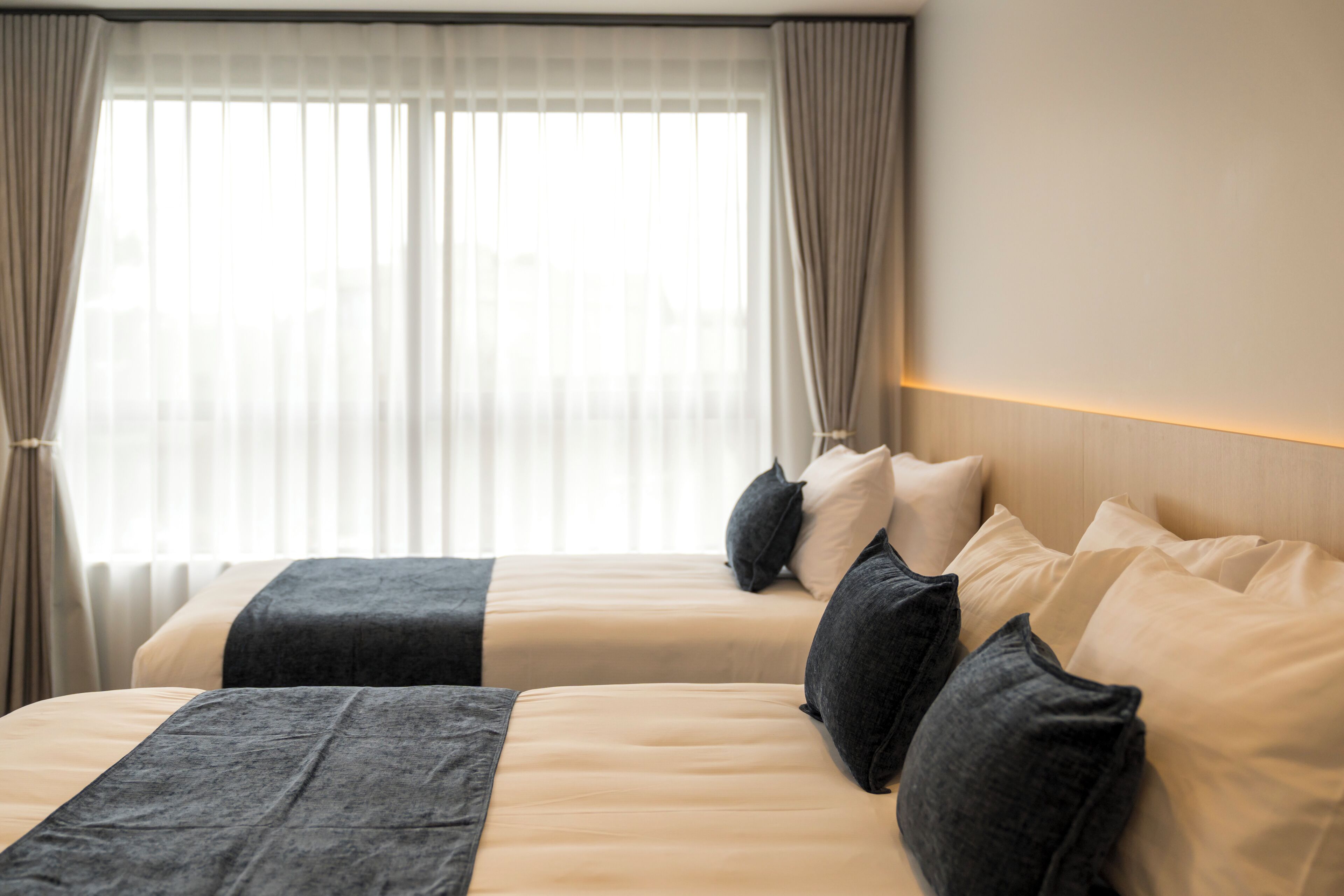 Una camera d'albergo con due letti, biancheria beige, coperte grigio scuro e un'ampia finestra con tende leggere.