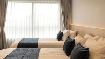 Una camera d'albergo con due letti, biancheria beige, coperte grigio scuro e un'ampia finestra con tende leggere.