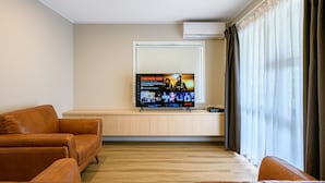 A modern living room with a flat-screen TV displaying a movie, leather sofas, a wooden cabinet, and a large window with curtains. - Takapuna International Motor Lodge (Auckland)