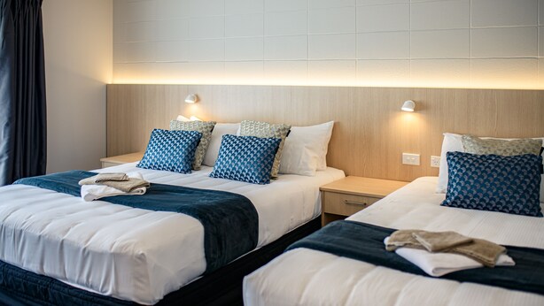 A hotel room with two beds, each with a white and dark blue bedspread, and matching pillows. There are bedside tables with lamps and a wall-mounted light fixture. - Takapuna International Motor Lodge (Auckland)