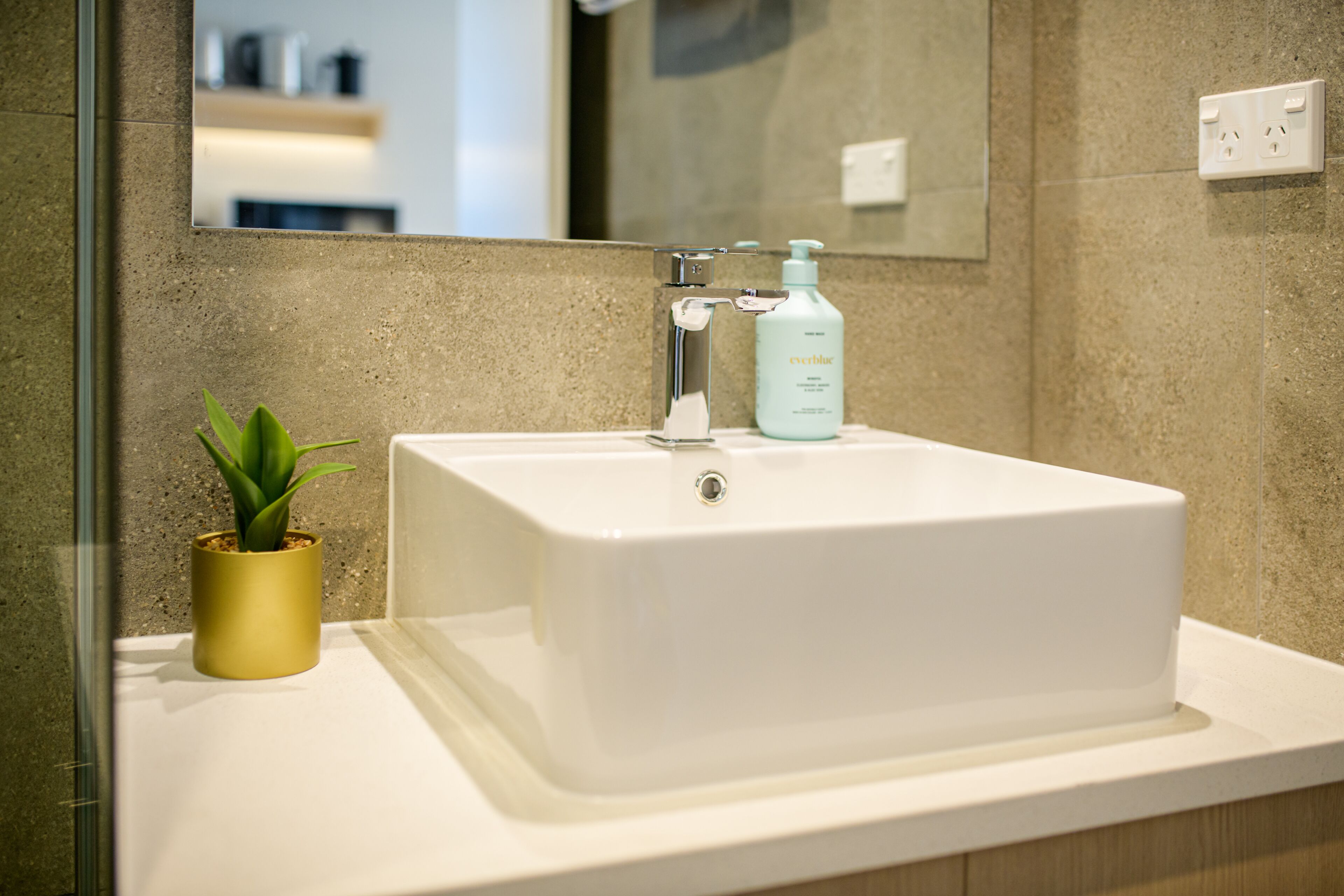 Une salle de bain avec un lavabo blanc, un vase doré orné d’une plante et un miroir au-dessus du lavabo.