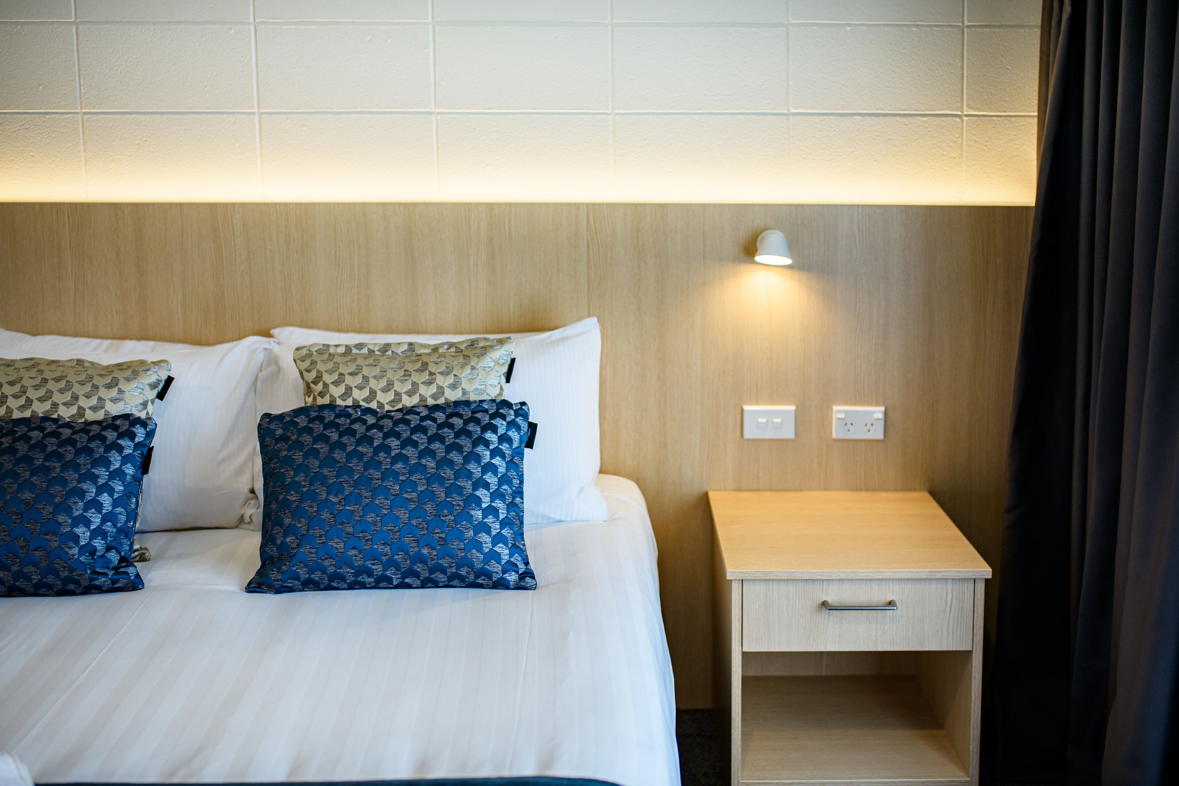 A hotel room with a neatly made bed, two pillows, a bedside table with a lamp, and a power outlet.