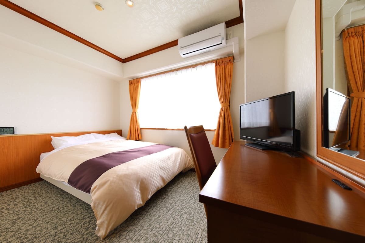 A hotel room with a bed, a desk with a television, an air conditioning unit, and a window with curtains.
