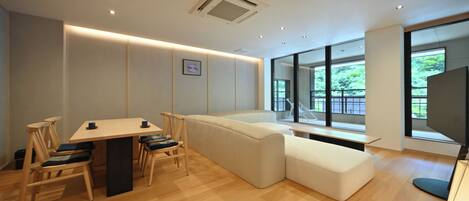 A modern interior with a wooden dining table, chairs, a sofa, and a large window offering a view of greenery.