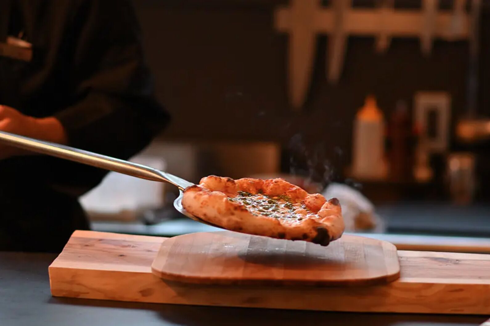 Een pizza wordt met een tang op een houten plank gelegd.