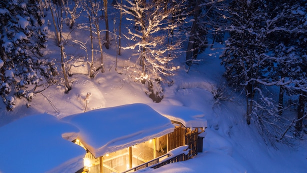 A snow-covered wooden cabin with a lit interior, surrounded by trees. - Hinanoyado Chitose (Tokamachi)