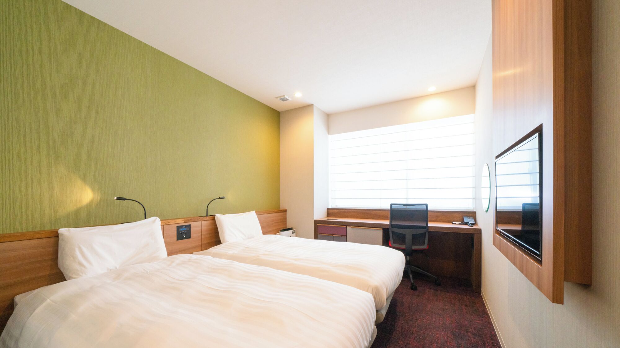A hotel room with two beds, a desk, a chair, a window with blinds, and a mounted TV.