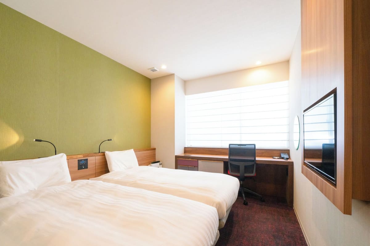 Ett hotellrum med två sängar, ett skrivbord, en stol, ett fönster med persienner och en väggmonterad TV.
