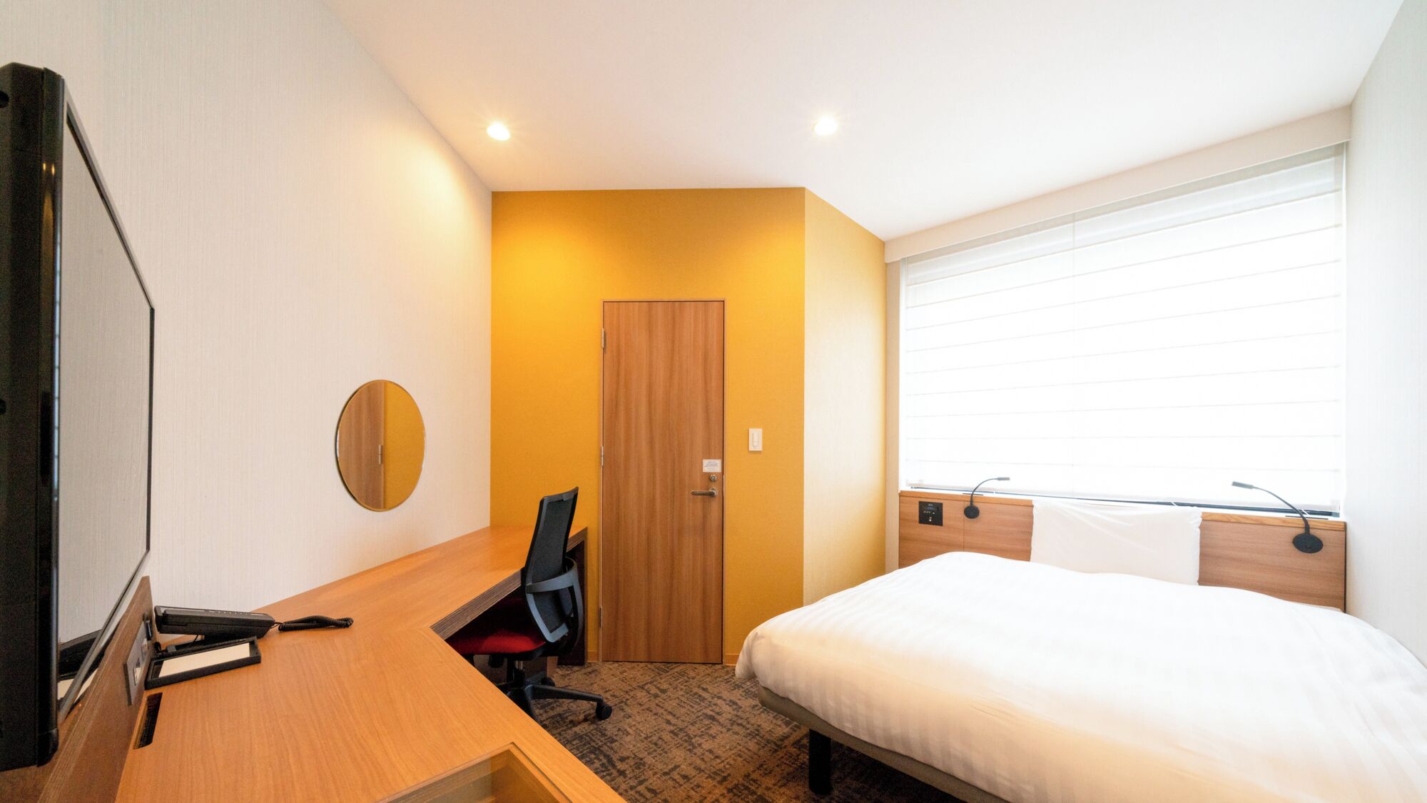 A hotel room with a bed, a desk with a chair, a round mirror, and a door.