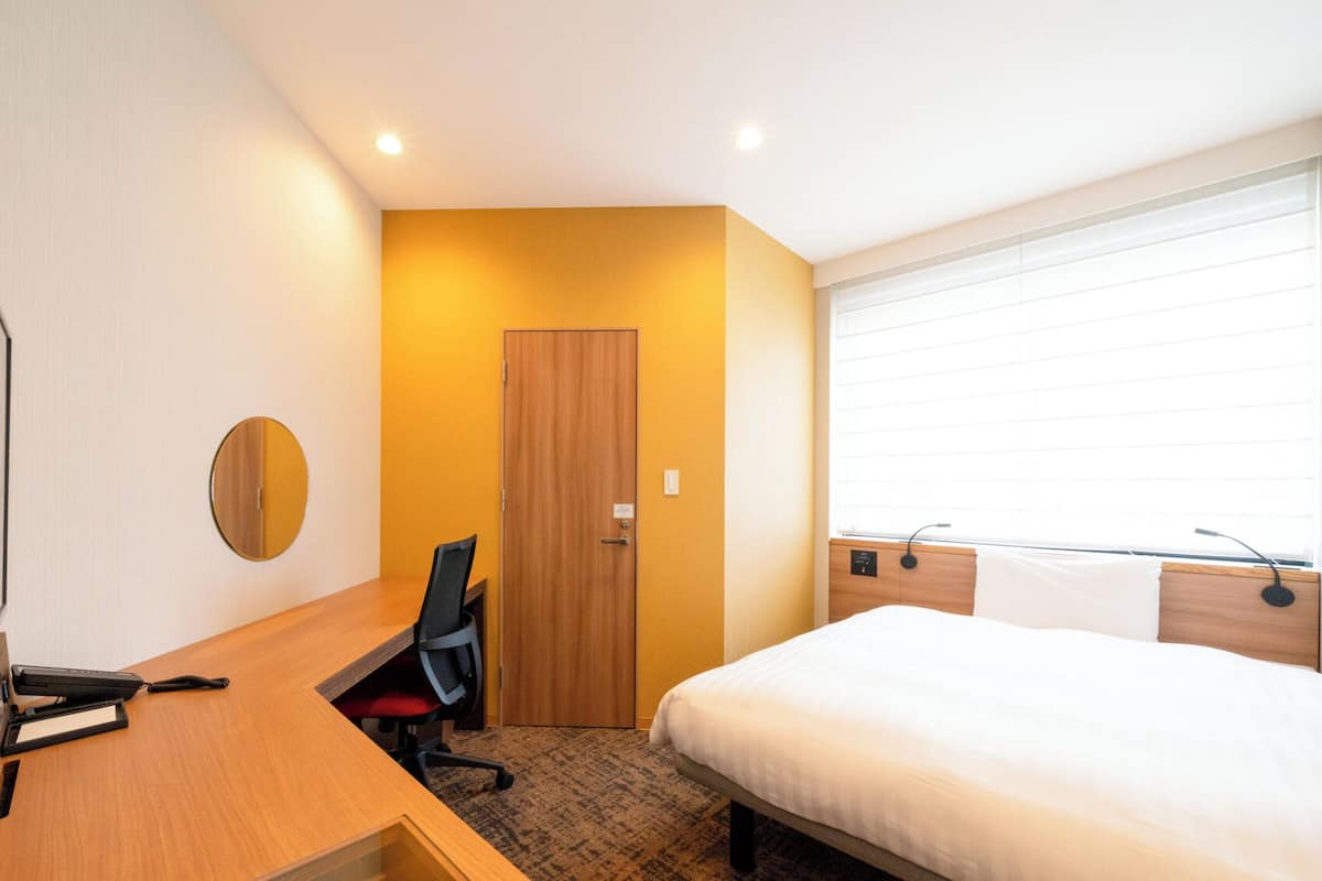 Ett hotellrum med en säng, ett skrivbord med en stol, en rund spegel och en dörr.