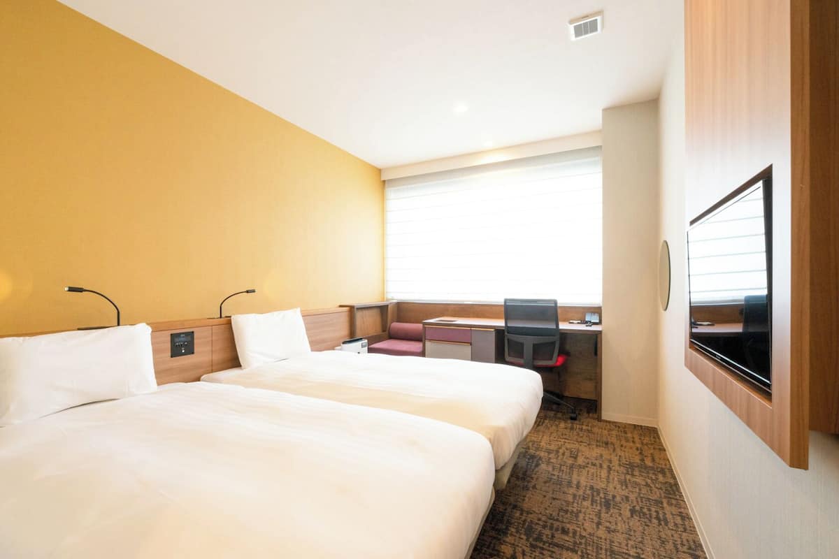 Ett hotellrum med två sängar, ett skrivbord, en stol, en tv och ett fönster med persienner.