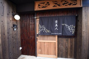 Una entrada japonesa tradicional con un portón de madera, una valla de bambú y un estandarte negro con caligrafía dorada.