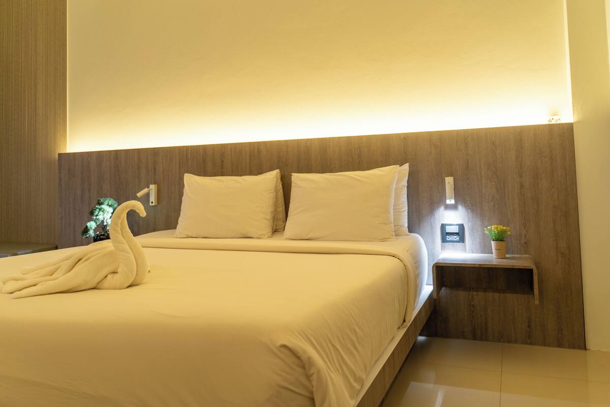 A neatly made bed with a swan-shaped towel, a bedside table with a plant, and a wall-mounted light.