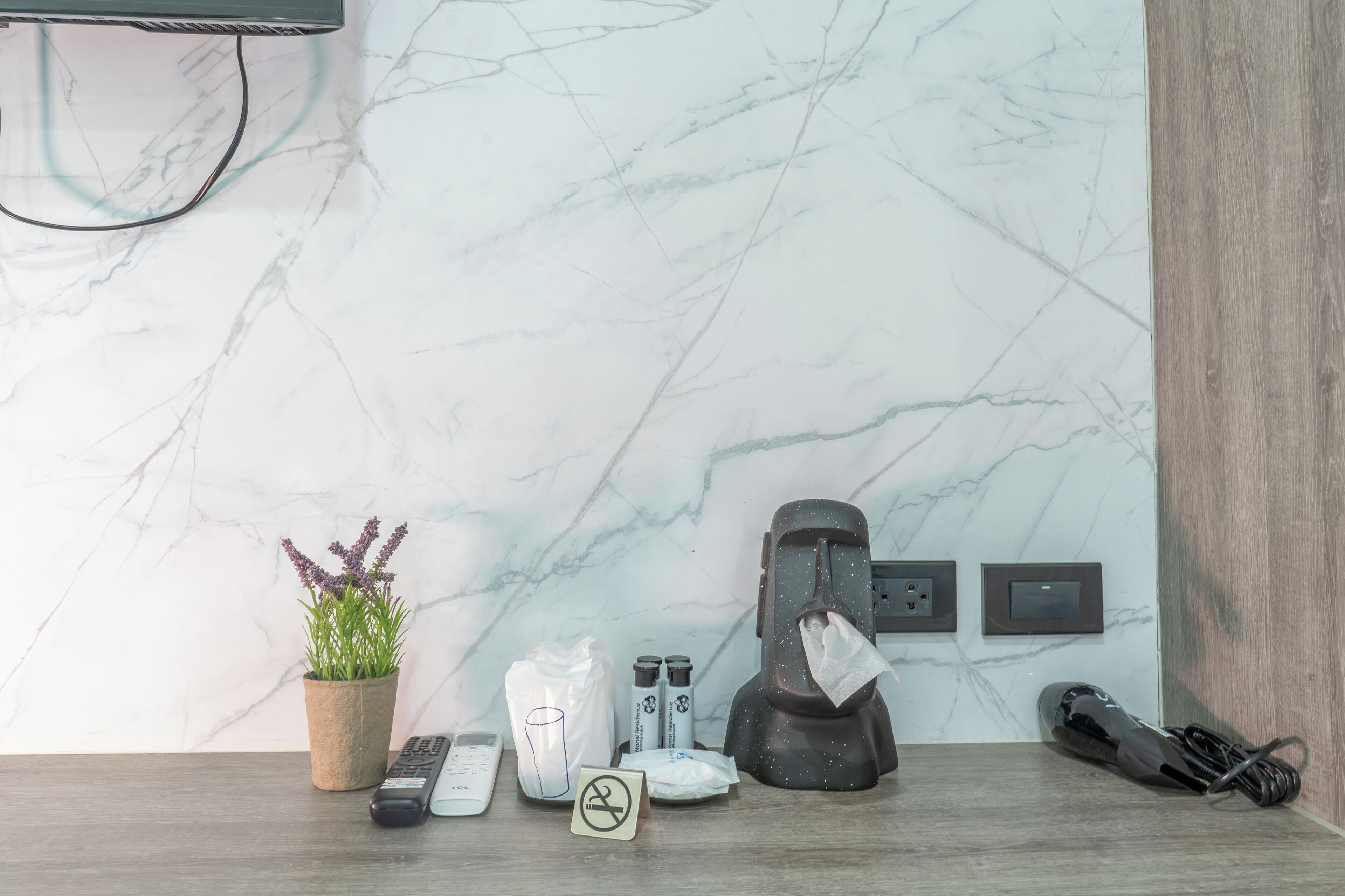 A bathroom with a marble wall, a potted plant, a towel dispenser, a trash bin, and a hairdryer on the floor.
