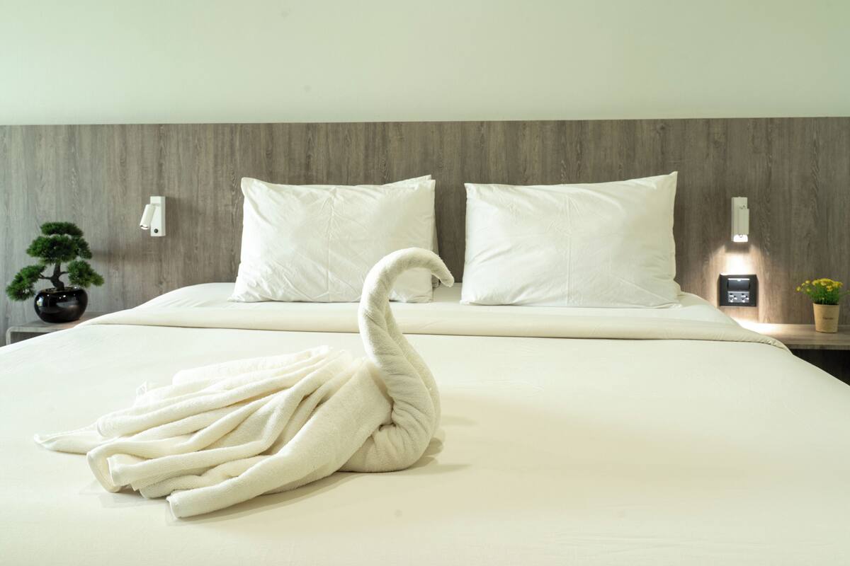 A neatly made bed with a swan-shaped towel, two pillows, a bedside table with a lamp, and a small potted plant.