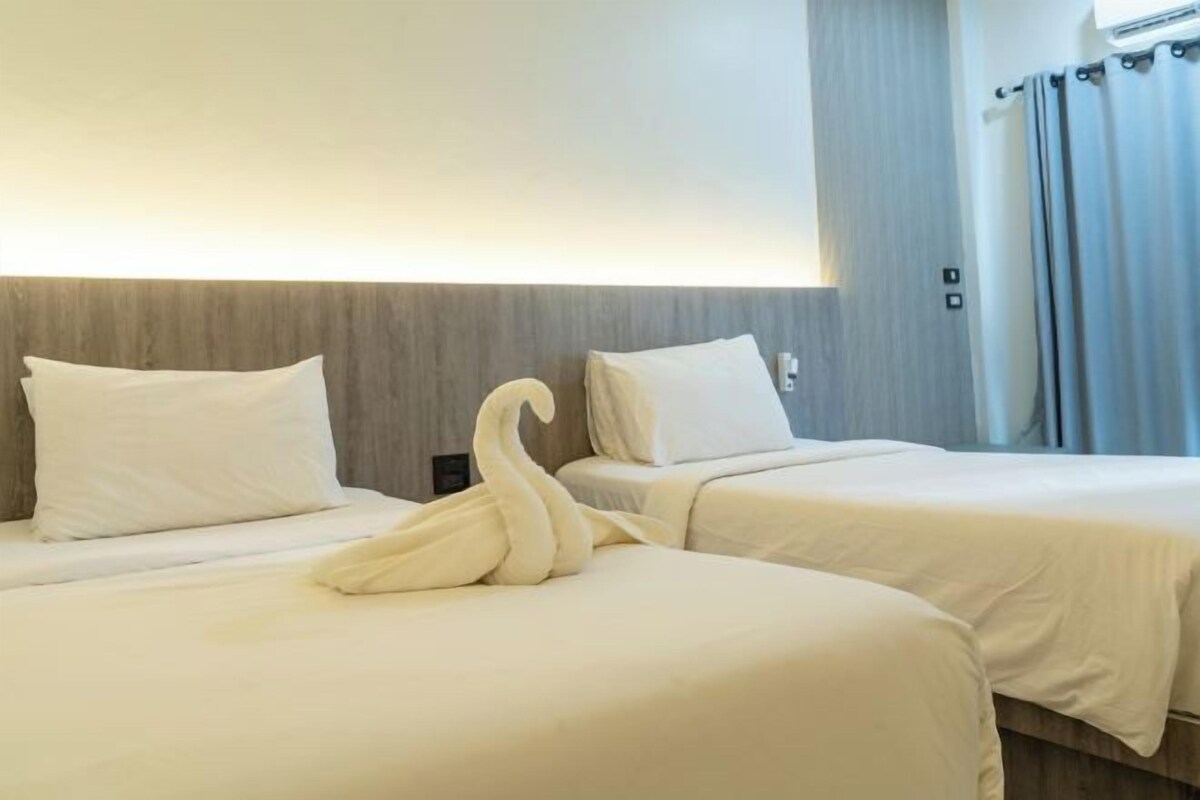A hotel room with two beds, each with white bedding and pillows. There is a swan-shaped towel on the bed in the foreground. The room has a wooden headboard and a curtain on the window.