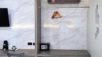 Una habitación moderna con pared de mármol, un escritorio con silla, un televisor montado en la pared, un estante con una bolsa y un perchero, y una caja fuerte pequeña.