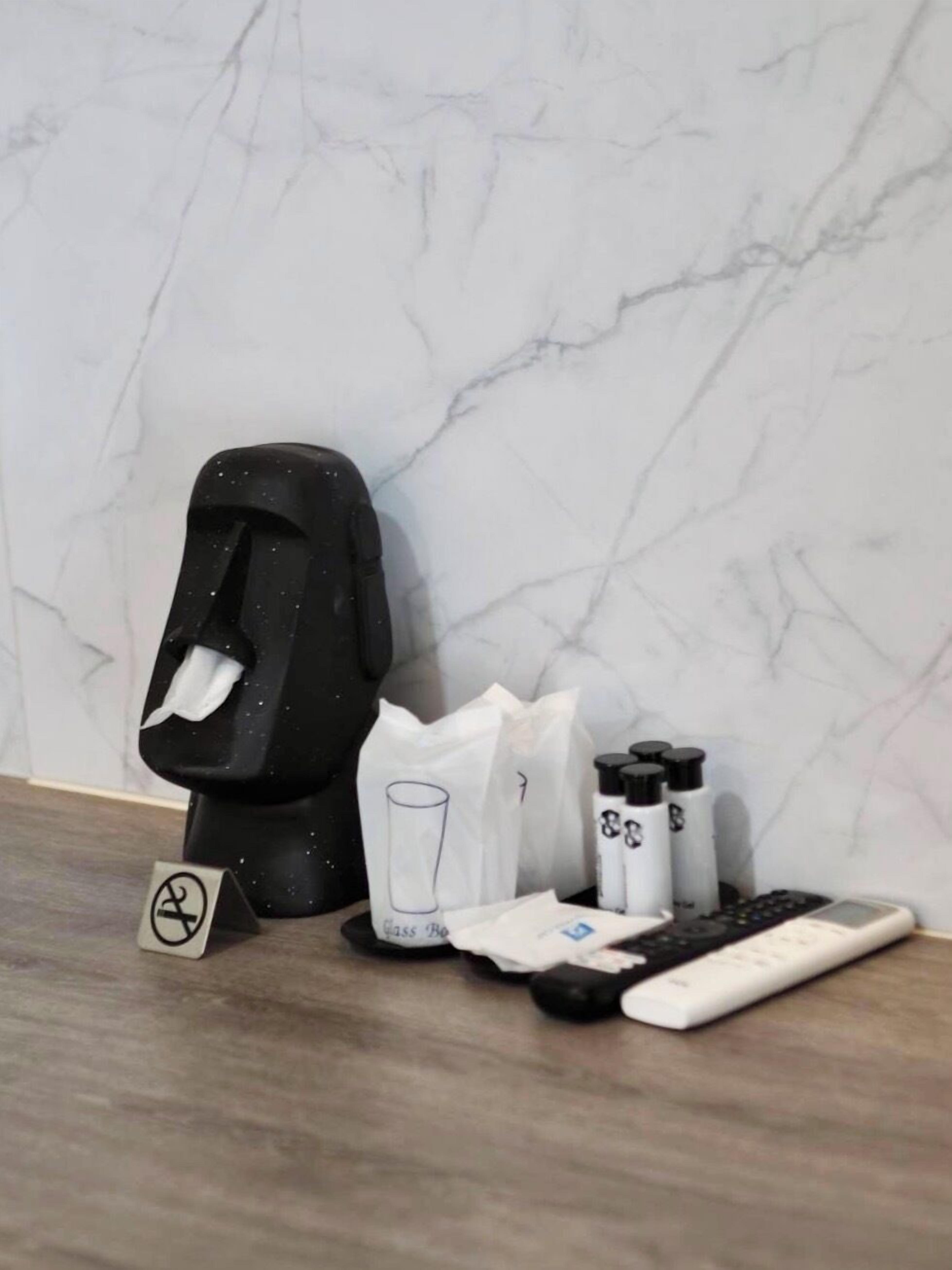 A bathroom with a marble wall, a black trash bin, a glass bottle, a white bag, and a remote control.