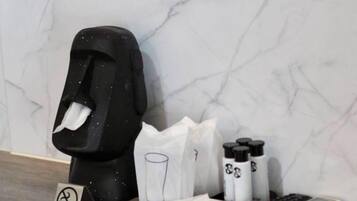 A bathroom with a marble wall, a black trash bin, a glass bottle, a white bag, and a remote control.