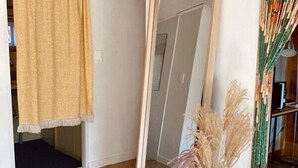 Una habitación con piso de madera, un espejo grande, un tapete estampado, un jarrón con plantas secas y una cortina amarilla.