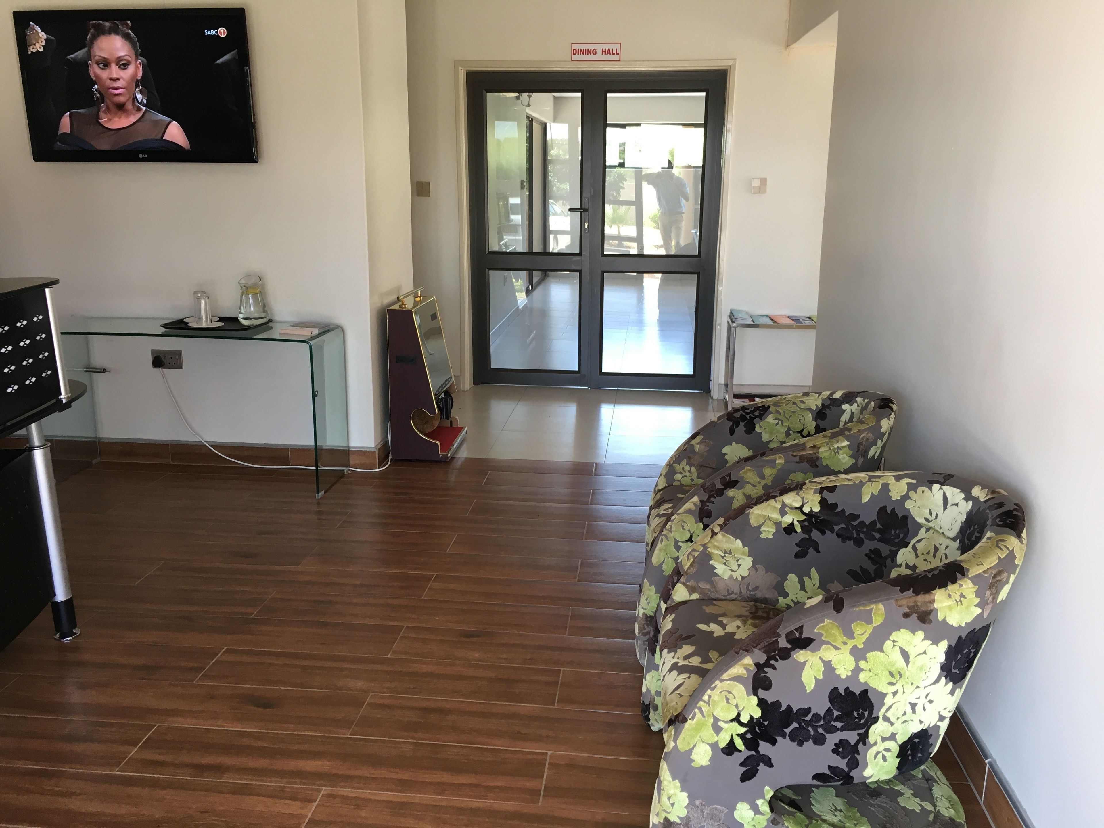 Una habitación con piso de madera, un sofá estampado con motivos florales, una mesa de vidrio con una cafetera y un televisor montado en la pared.