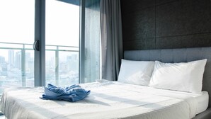 Une chambre d’hôtel avec un grand lit, une vue sur la ville grâce à une fenêtre du sol au plafond, et un lit parfaitement fait avec une serviette pliée.