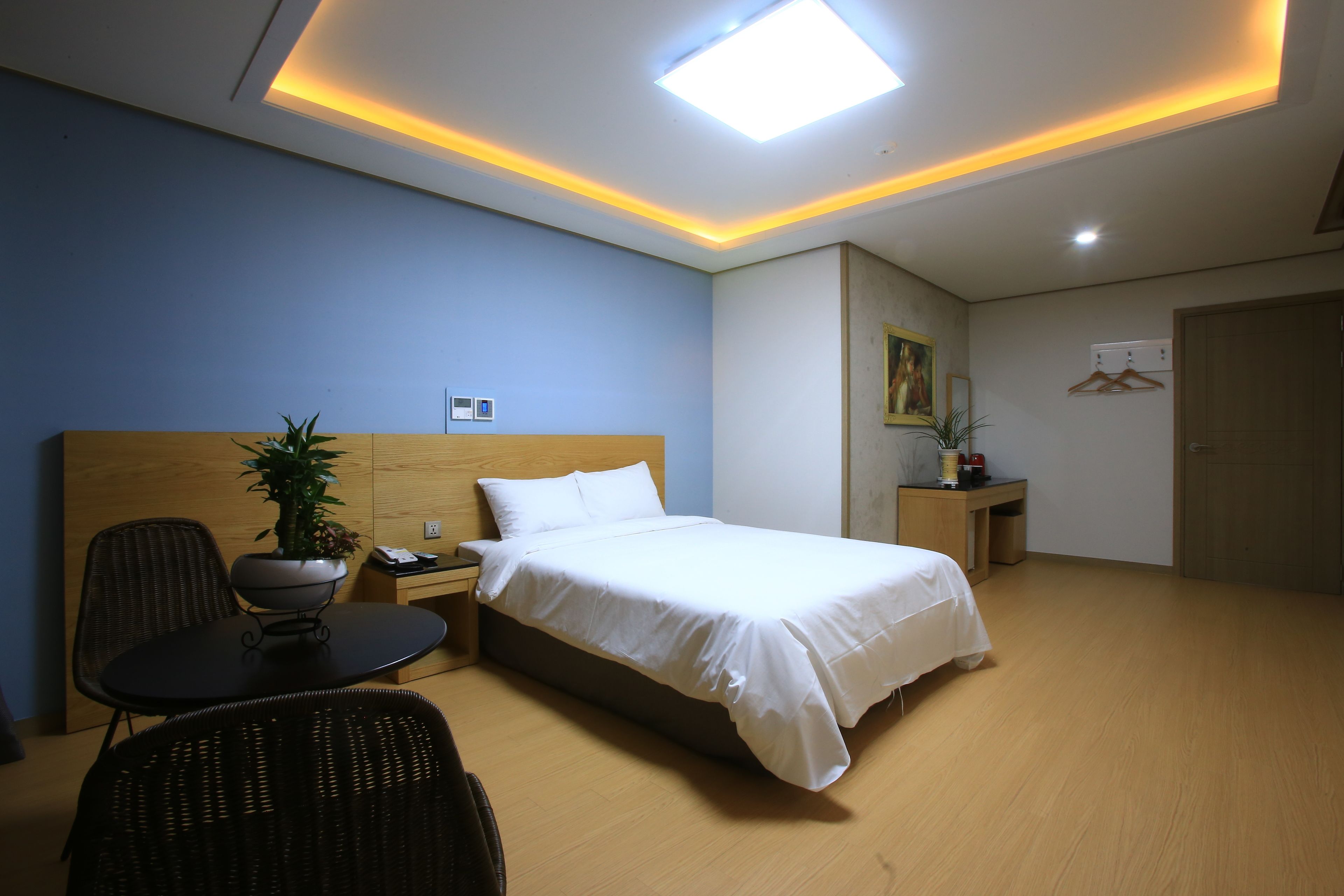 A hotel room with a bed, a desk, a chair, a potted plant, and a ceiling light.