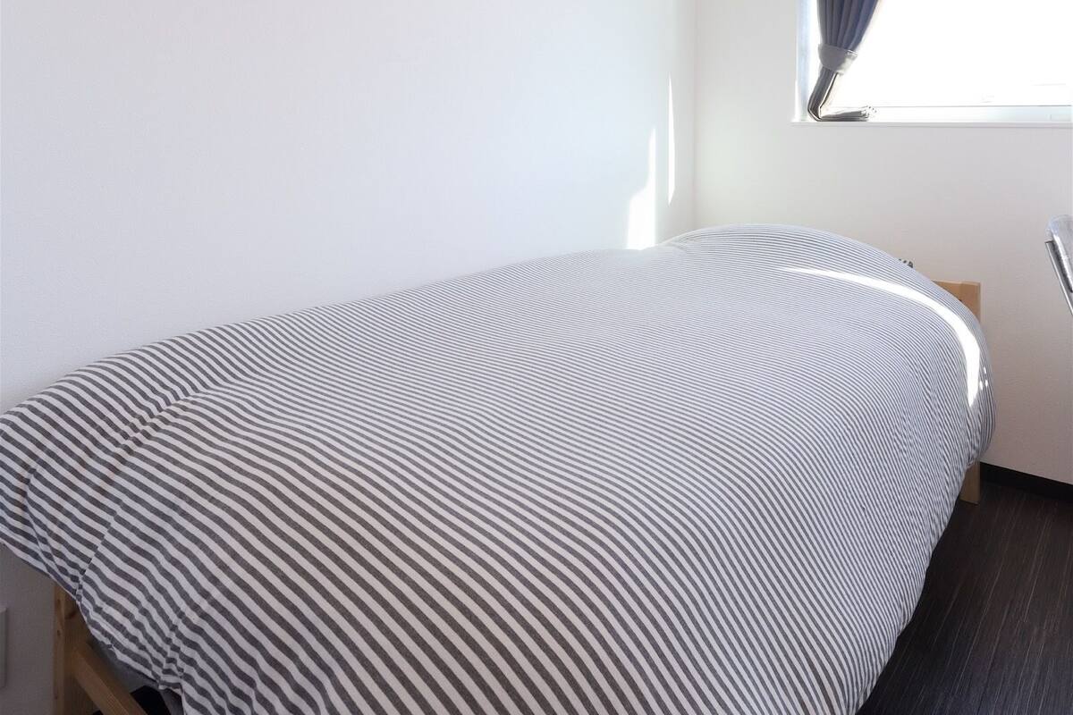Une petite chambre à coucher, bien rangée, avec un lit simple, une fenêtre avec des rideaux et un luminaire au plafond.