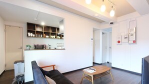 A modern living room with a sofa, coffee table, and kitchen area with shelves and appliances. - Shonan-Enoshima Seaside Guest House – Hostel (Fujisawa)
