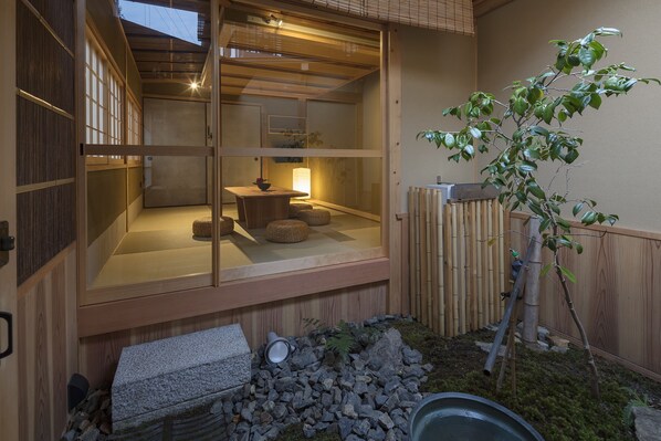 A traditional Japanese-style room with tatami flooring, a low wooden table with cushions, and a sliding glass door leading to a garden area with a small tree and rocks.