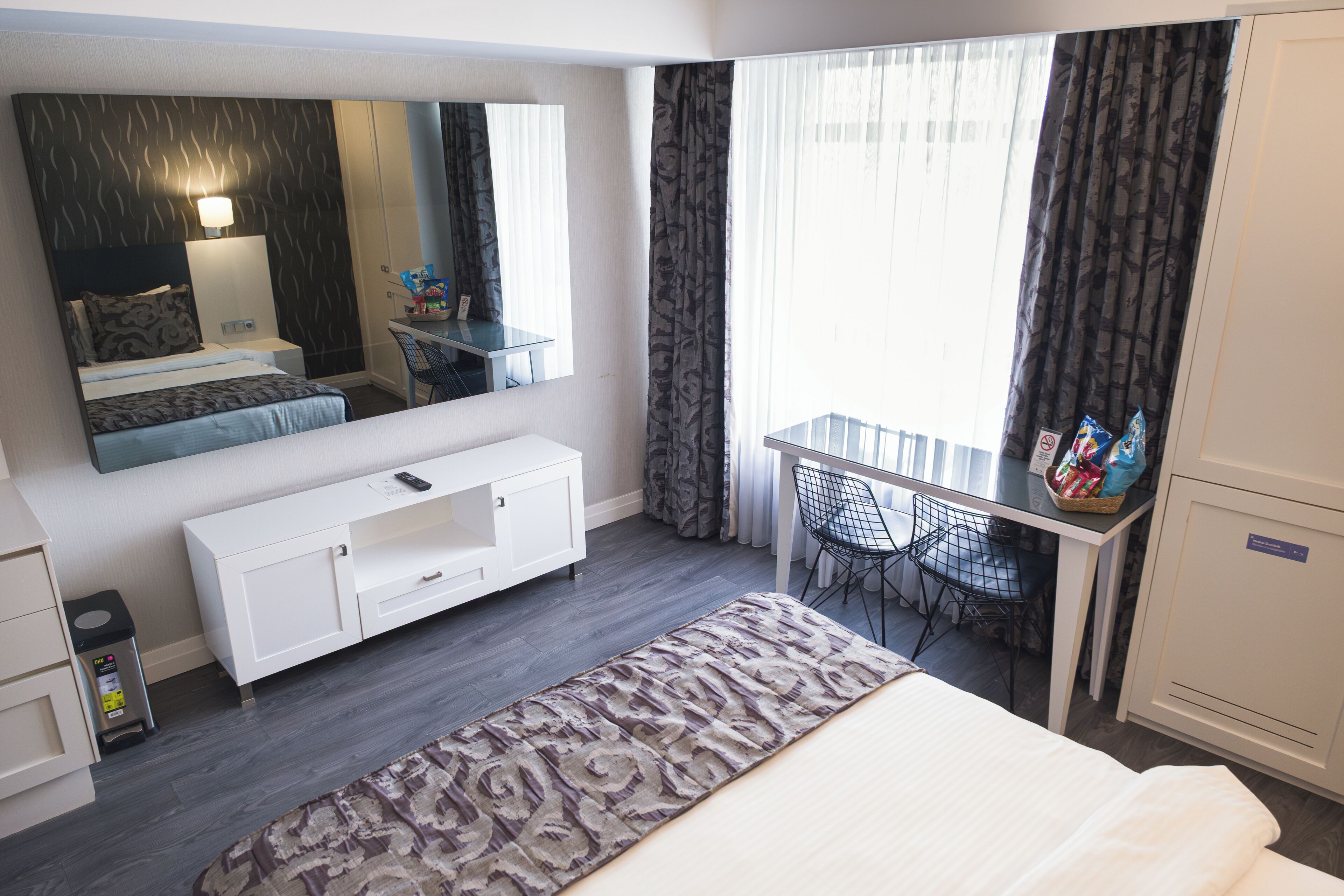 A hotel room with a bed, a desk with a chair, a small table, a mirror, and a wardrobe.