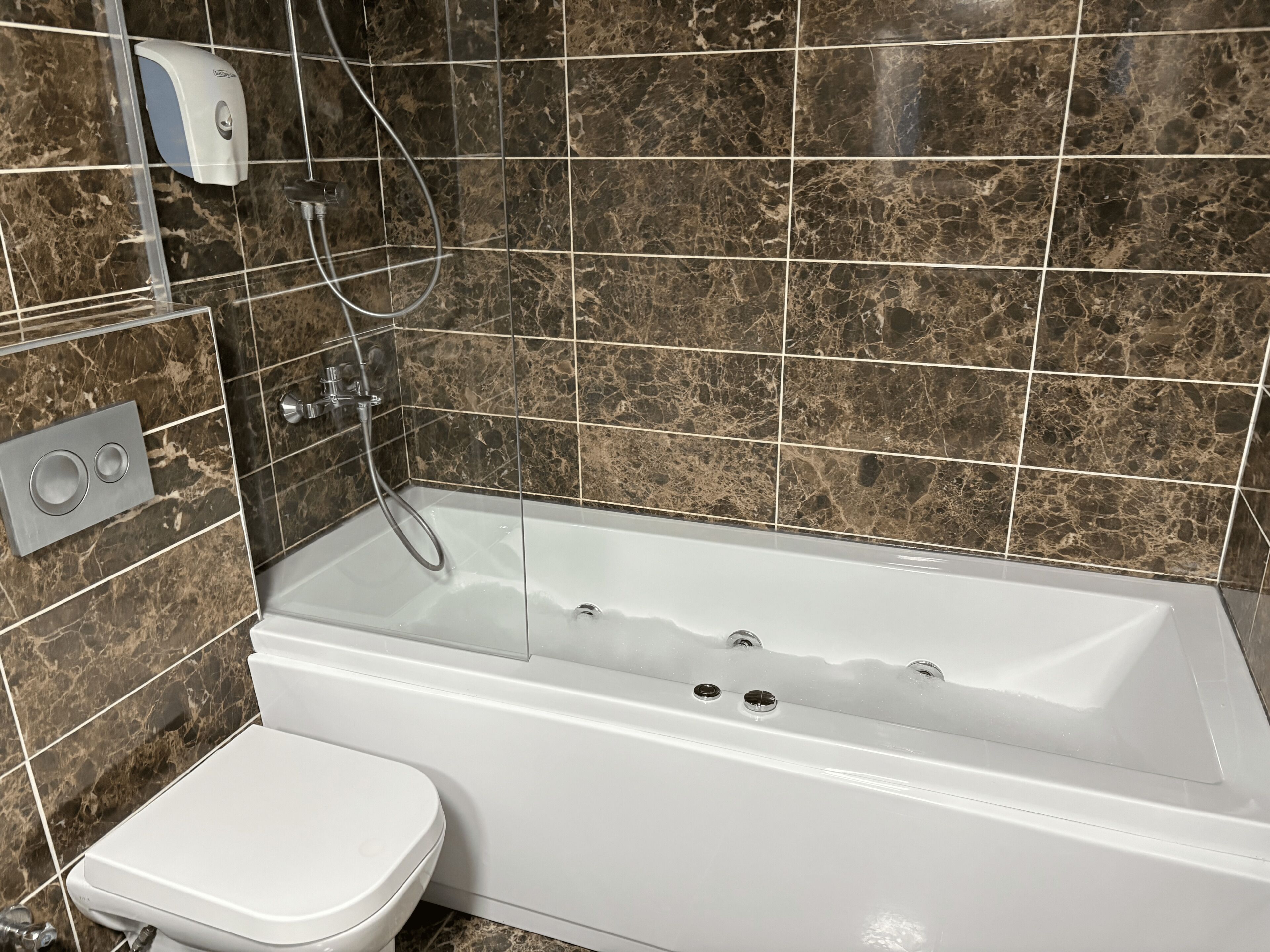 A bathroom with a white bathtub, a toilet, and a hand soap dispenser on the wall.