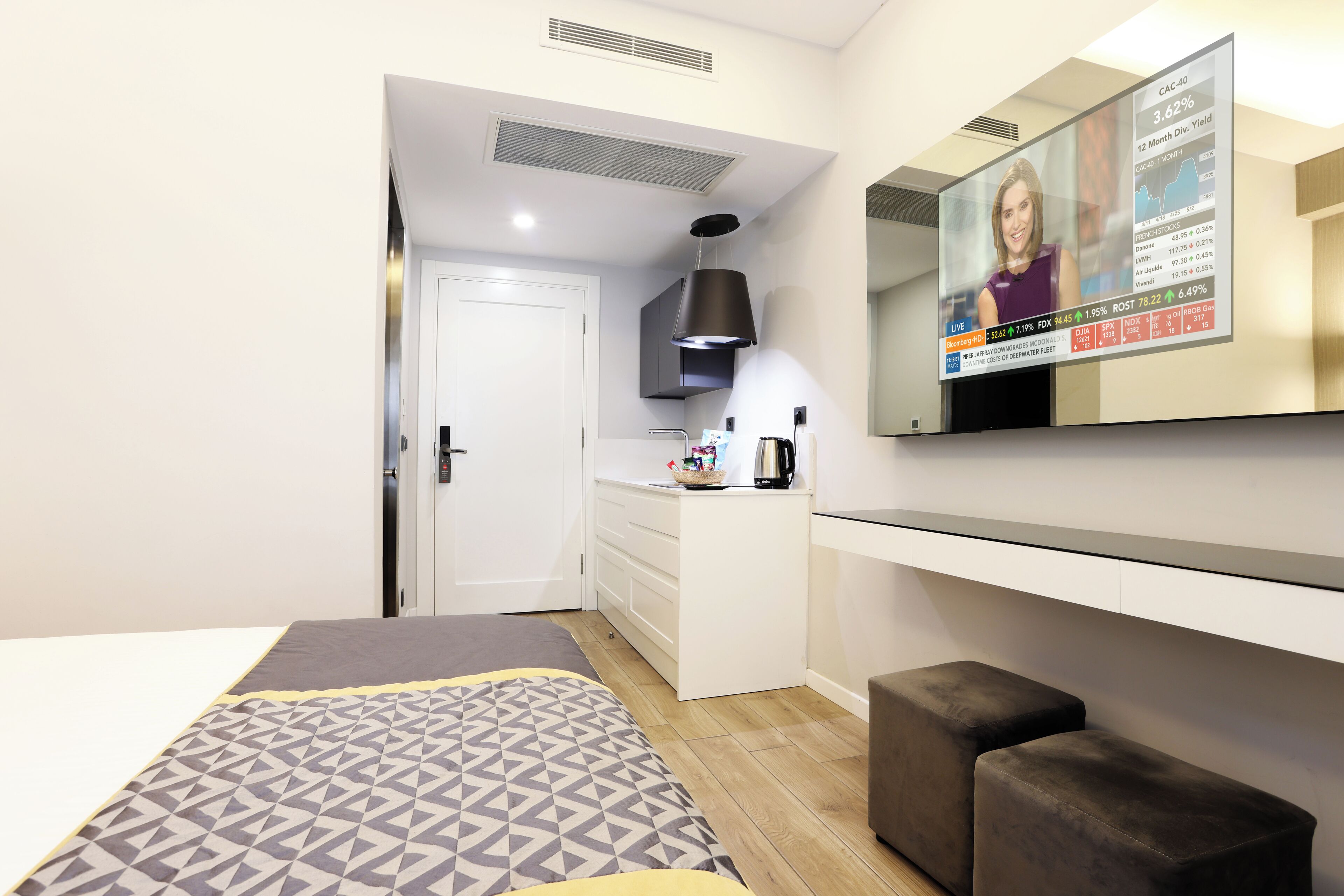 A modern hotel room with a bed, a TV mounted on the wall, a kitchenette, and a small seating area.