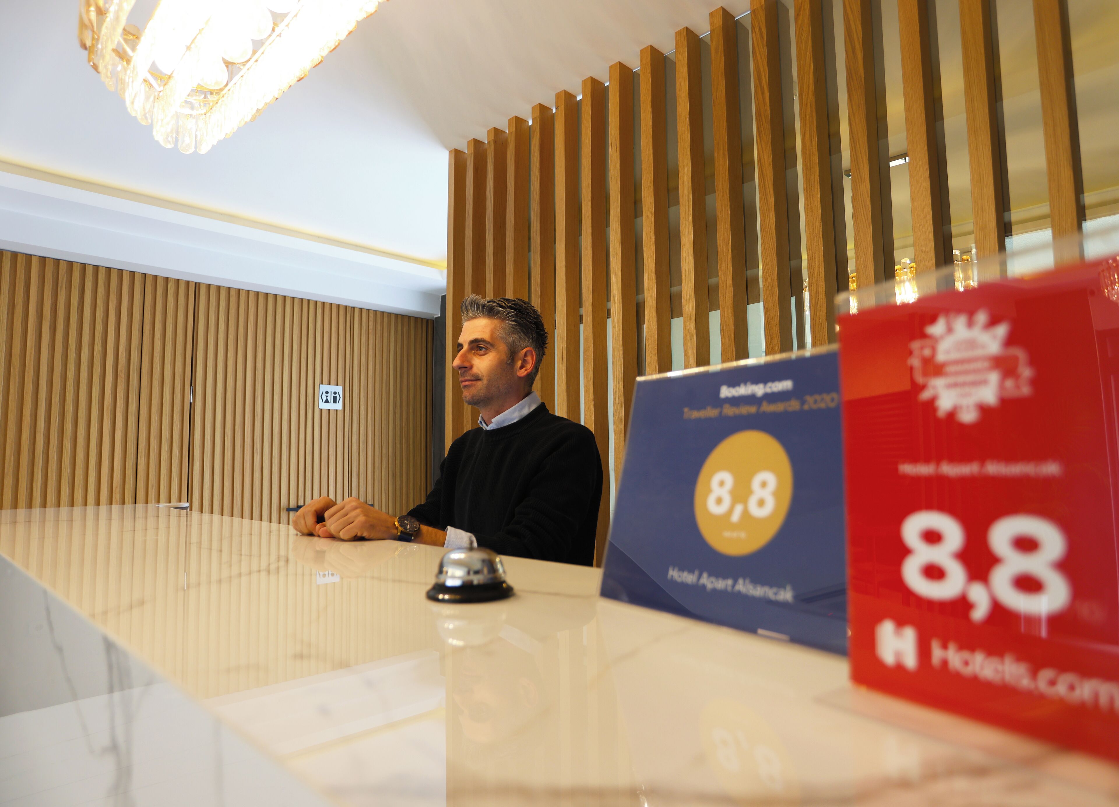 Una recepción de hotel con pared de paneles de madera y una lámpara de araña. Hay un cartel que indica una calificación de 8.8 de Booking.com.