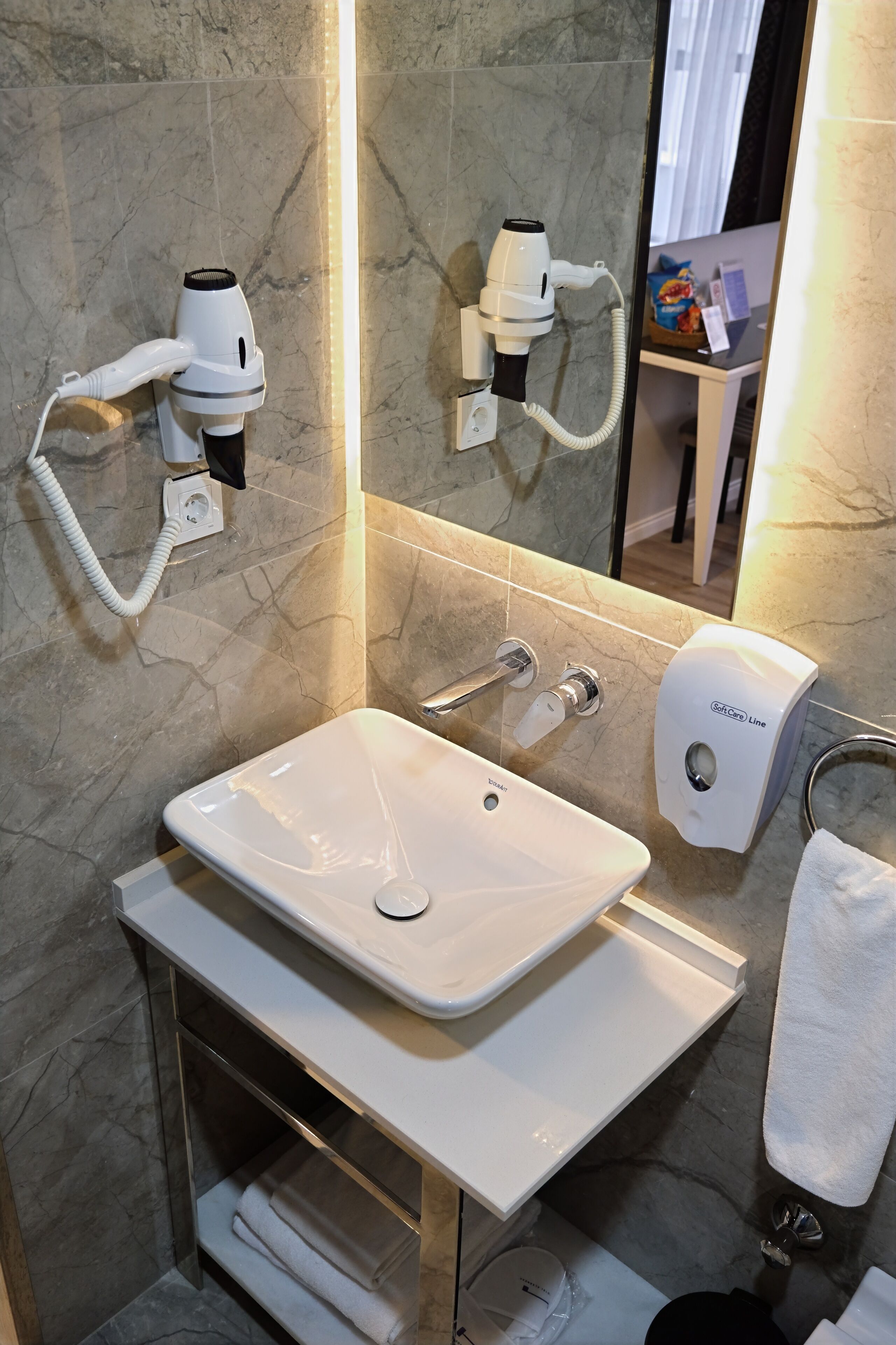 Bagno con lavandino bianco, asciugacapelli portatile, portasciugamani e dispenser per sapone.