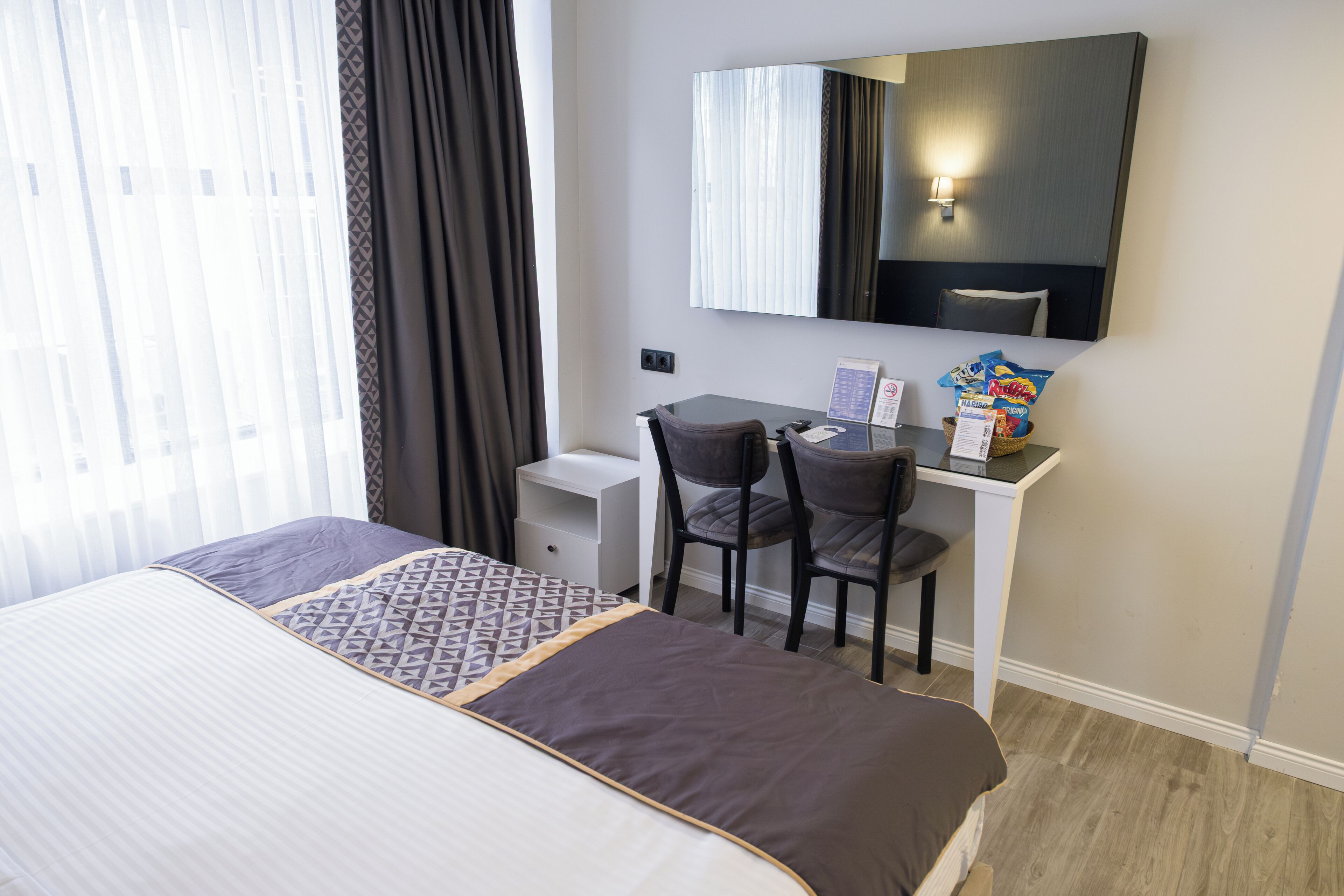 A hotel room with a bed, a desk, two chairs, a mirror, and a window with curtains.