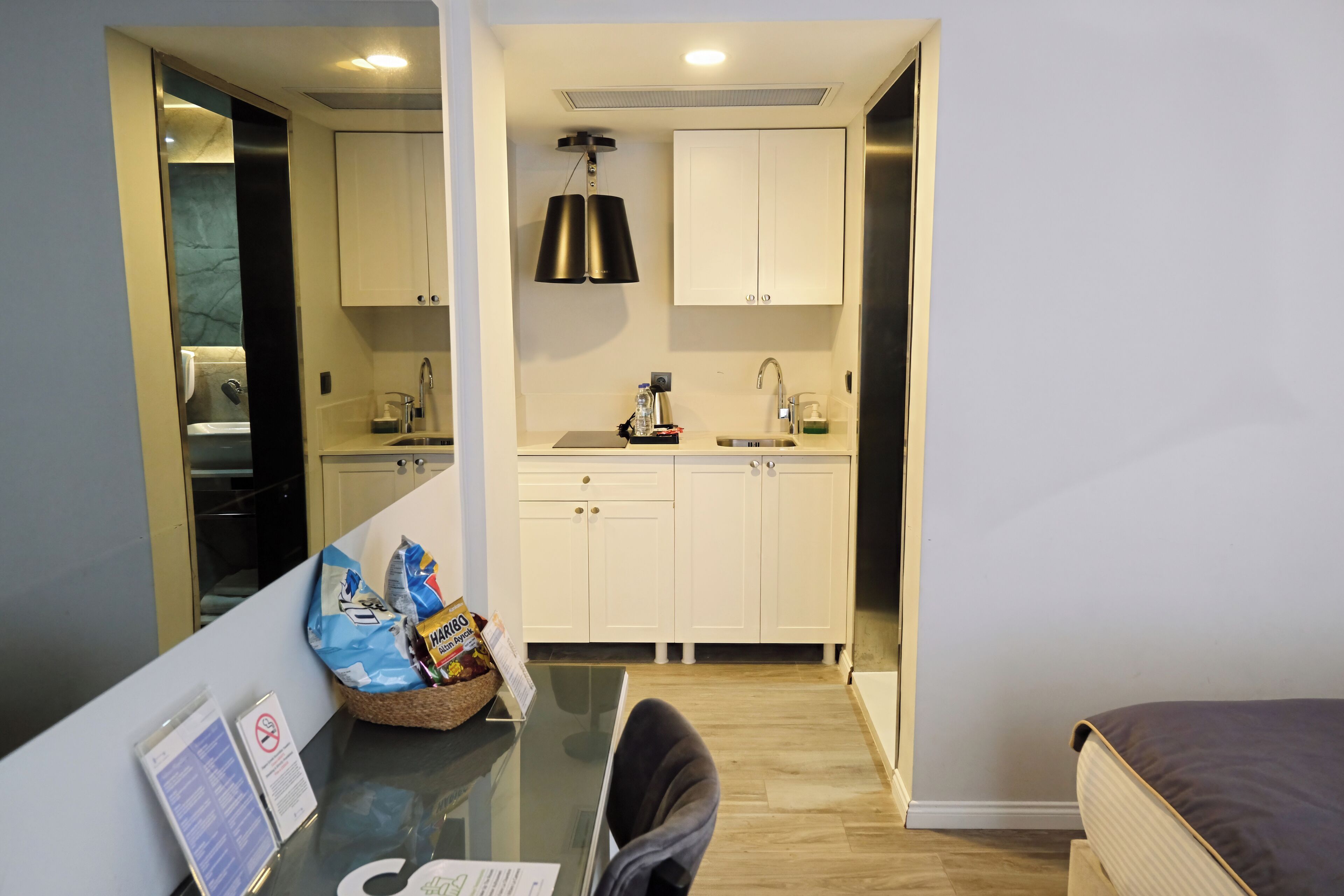 A compact hotel room with a kitchenette, a glass table, a chair, and a bed.