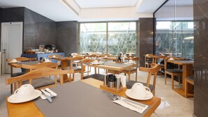A modern dining area with wooden tables and chairs, a coffee station, and large windows offering a view of greenery outside. - Riva Hotel Alsancak (Izmir)