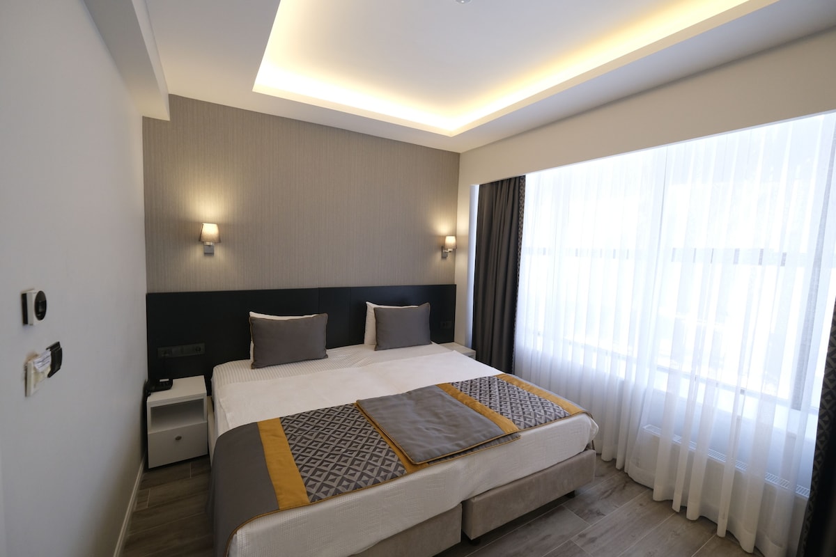 A modern hotel room with a large bed, bedside tables, a headboard, and a large window with sheer curtains.