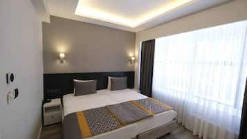 Una moderna camera d'albergo con un ampio letto, comodini, una testiera e una grande finestra con tende leggere.
