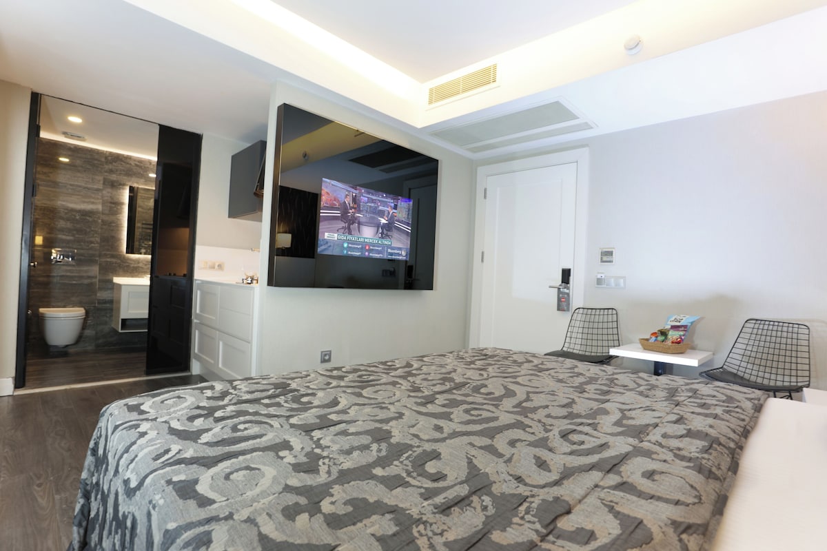 A modern hotel room with a large bed, a TV mounted on the wall, a small table with a basket, and a door leading to a bathroom.