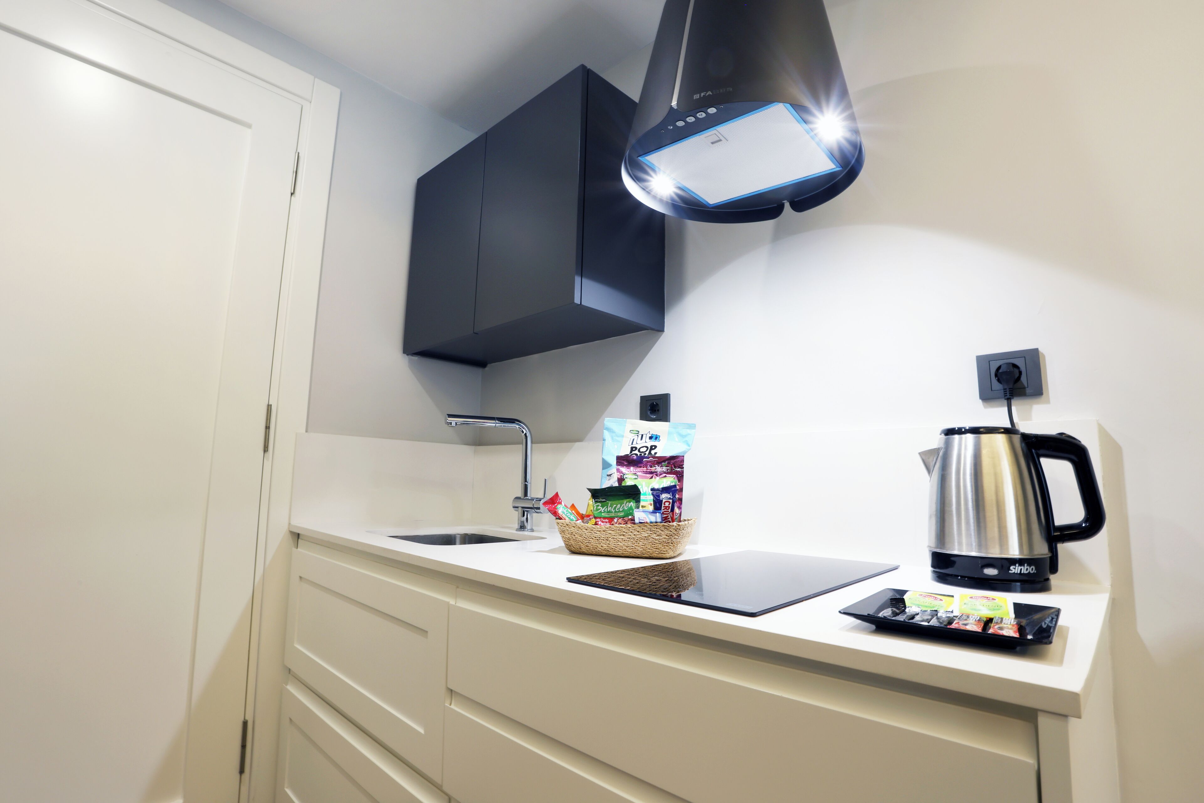 A modern kitchen with a white countertop, a black range hood, a stainless steel kettle, and a basket of snacks.
