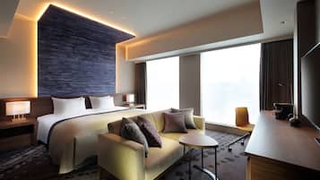 A modern hotel room with a large bed, a sofa, a small round table, and a desk.