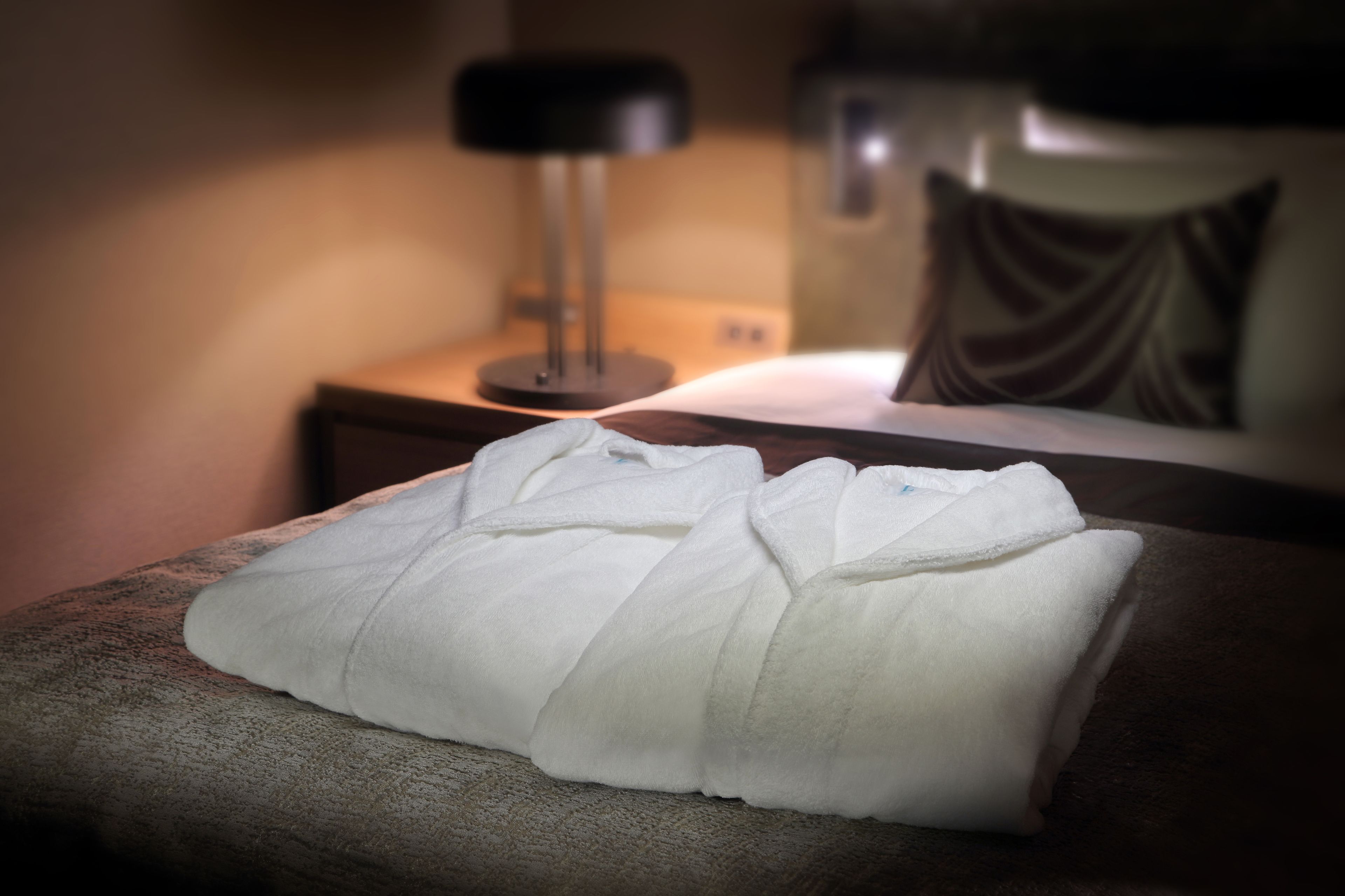 A neatly folded white towel on a textured brown chair in a dimly lit room with a lamp and a pillow on the bed.