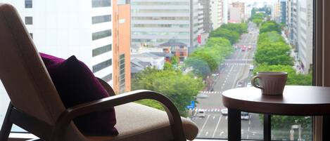Una habitación de hotel con vista a una calle de la ciudad, una silla con cojín y una mesa con una taza.