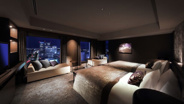 A modern hotel room with a large bed, a sofa, and a TV. The room has a city view through large windows.