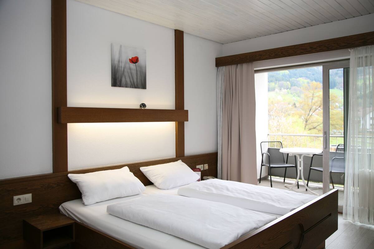 A hotel room with a large bed, a small table and chairs, a painting on the wall, and a balcony with a view of trees.